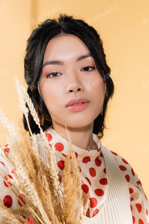 Preview: Portrait of trendy asian woman looking at camera near blurred spikelets on orange background
