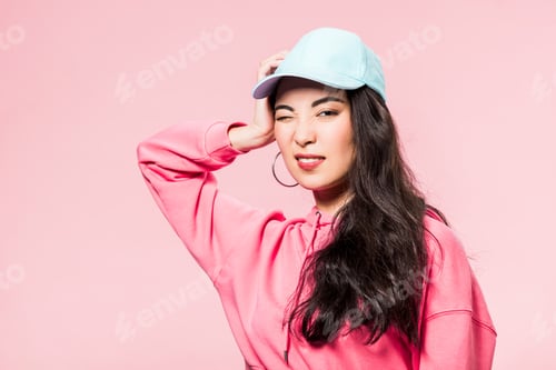 Preview: attractive asian woman in pink pullover and cap winking isolated on pink