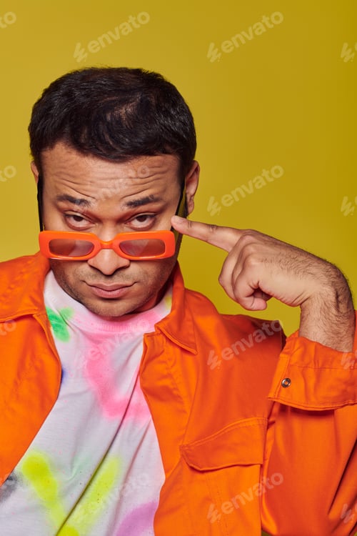 Preview: self-expression, confident indian man adjusting orange sunglasses on yellow backdrop, style icon