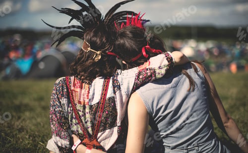Preview: Rear view of two young women at a summer music festival wearing feather headdresses, arm around