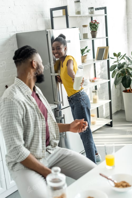 Preview: smiling african american girlfriend giving bottle of milk to boyfriend at kitchen