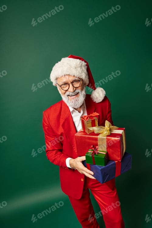 Preview: handsome cheerful Santa posing with pile of gifts in hands on green backdrop, Christmas concept