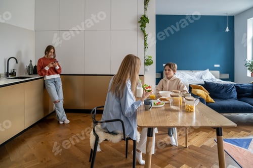 Preview: Teen girl using smartphone standing away of family having breakfast at kitchen table
