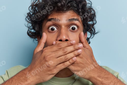 Preview: Close up shot of stupefied curly haired Hindu man covers mouth with both hands stares bugged eyes