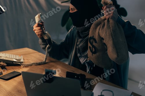 Preview: Angry terrorist in black mask holding money bags and looking at handset