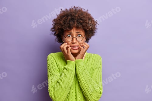 Preview: Photo of dark skinned worried woman bites finger nails and looks anxious at camera worries before im