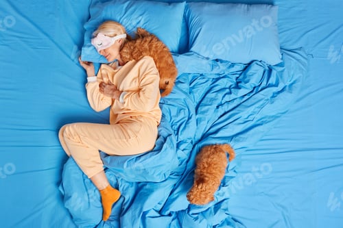 Preview: Relaxed blonde adult woman wears blindfold on eyes sleeps well in bed wears comfortable pajama two f