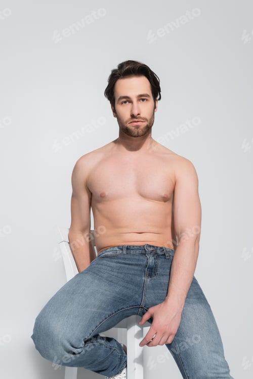 Preview: brunette shirtless man in jeans sitting on white chair and looking at camera on grey