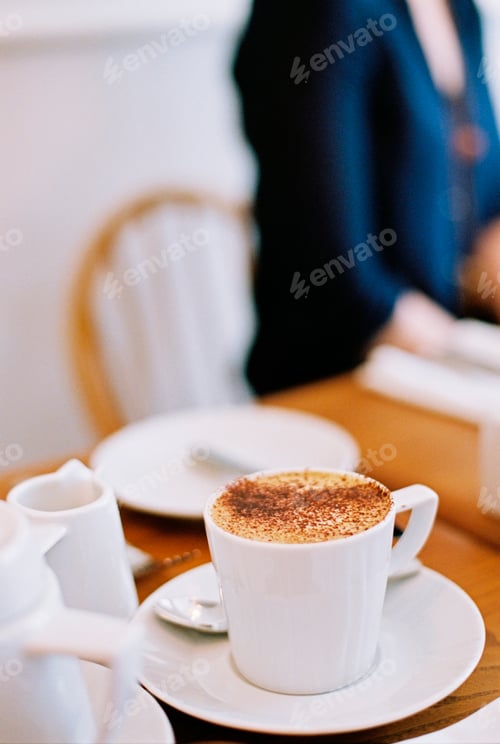 Preview: A woman seated at a table. A table setting and a large cup of coffee.