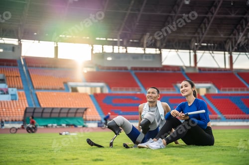 Preview: Asian para-athlete with prosthetic blades and trainer sit in stadium.