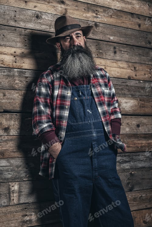Preview: Vintage Worker Man With Long Gray Beard In Jeans Dungarees. Standing In Front Of Wooden Wall.