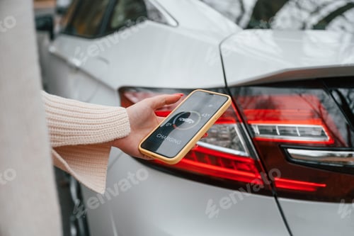 Preview: With smartphone in hand. Woman is standing near her electric car outdoors on the road
