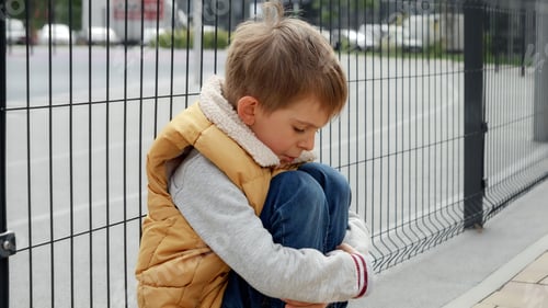 Preview: Portrait of little boy crying while sitting alone next to metal fence on street