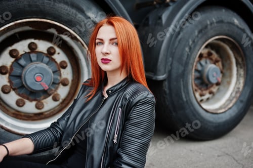 Preview: Red haired stylish girl wear in black, sitting against large truck wheels.