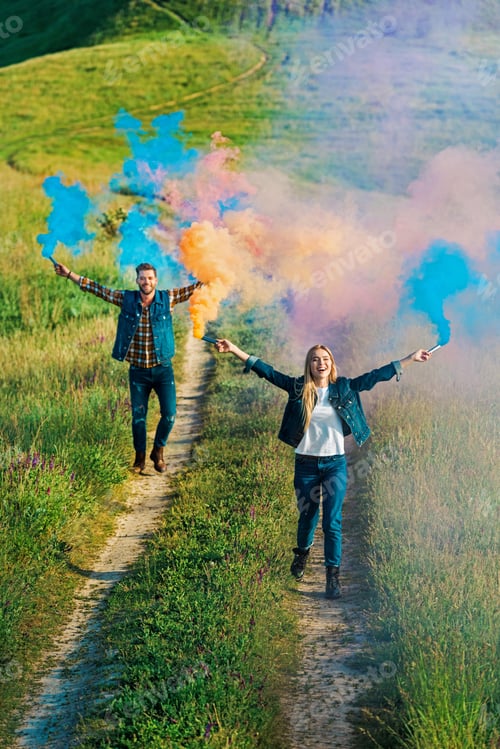 Preview: high angle view of young couple holding colorful smoke bombs on rural meadow