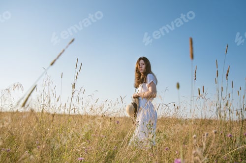 Preview: A young girl with glasses, a white summer dress with a straw hat in a field with flowers at sunset.