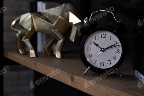 Preview: Black alarm-clock with bull statuette on wooden shelf.