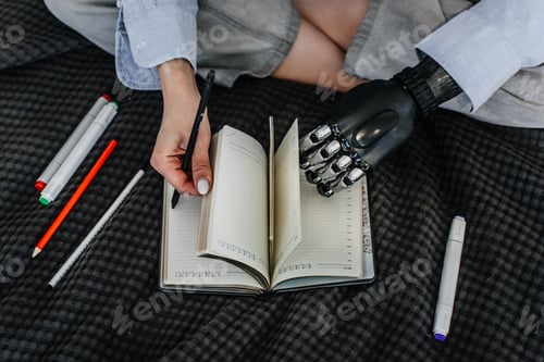 Preview: Cropped image of woman sitting on bed opens diary using biomechanical hand. Girl with artificial arm