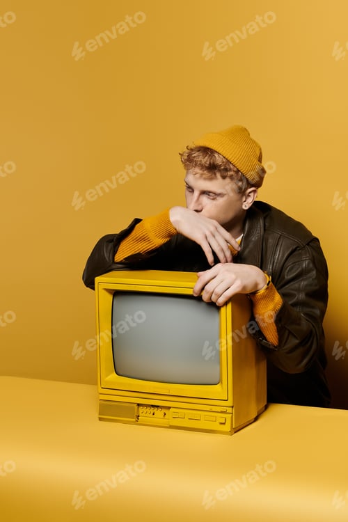 Preview: A stylish young man with red hair poses in a trendy winter outfit, seated with a vintage TV.
