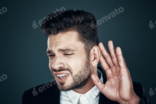 Preview: Charming young man in formalwear holding hand near ear while standing against grey background