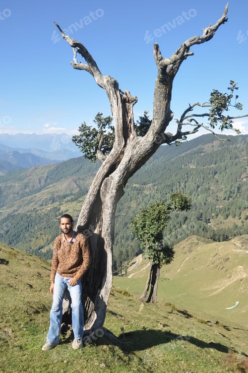 Preview: Full length of a stylish Indian young man looking at camera while posing with leaning against tree