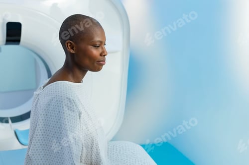 Preview: African American woman sitting on padded scanner table in radiology room wearing hospital gown