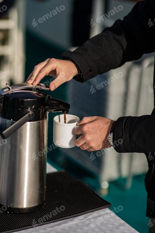 Preview: Person pouring freshly brewed coffee into a cup from a large stainless steel coffee pot