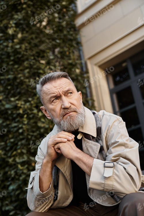 Preview: A fashionable senior man sits thoughtfully against a sleek wall, in trendy attire.