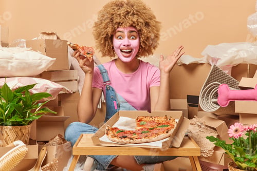 Preview: Cheerful amazed woman with curly hair eats appetizing pizza dressed in casual clothes relocates to n