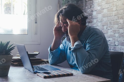 Preview: One adult man touching his head temples in front of a laptop computer in home office workplace