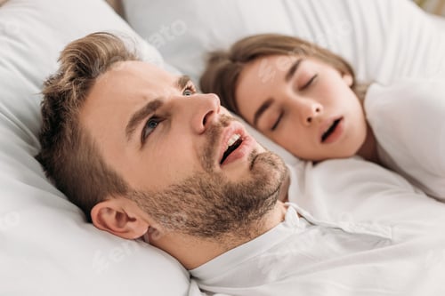 Preview: awakened man looking up while lying in bed near snoring wife