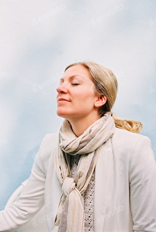 Preview: A woman with blonde hair sitting with her eyes closed feeling the sun on her face,