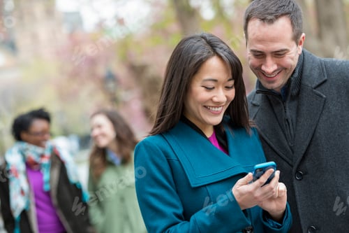 Preview: City park. A man and a woman in a coats looking at a smart phone.