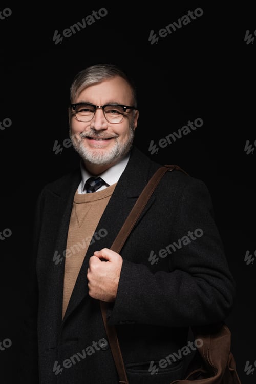 Preview: happy senior man in trendy coat and eyeglasses looking at camera isolated on black