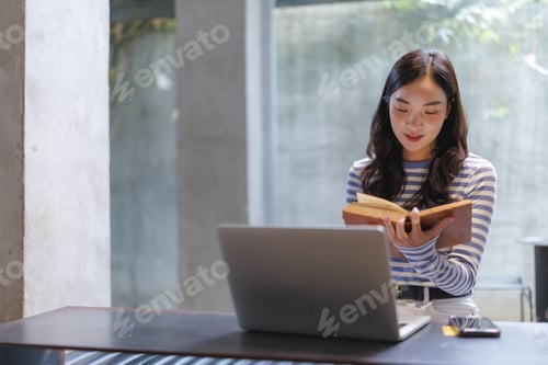 Preview: Young asian woman reading a book and studying with laptop in modern office space