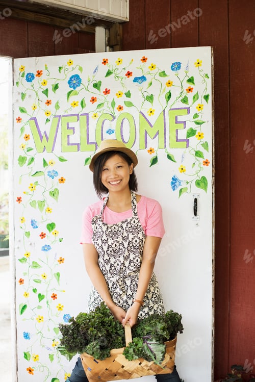 Preview: A young Asian woman by the Welcome sign at a farmstand with a large basket of vegetables