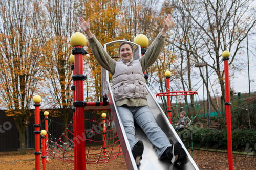 Preview: Happy mother sliding down playground slide with raised arms and smiling