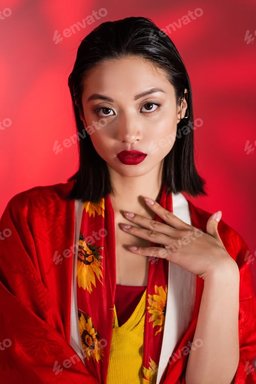 Preview: brunette asian woman in kimono cape and scarf with floral decor looking at camera on abstract red