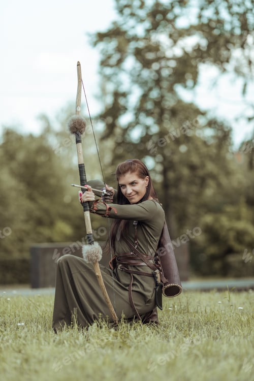 Preview: Young woman warrior with a bow sits in a clearing and aims from a bow, hunting in a green forest