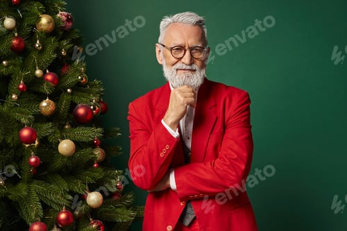 Preview: happy Santa in classy attire with glasses posing next to fir tree with hand on chin, winter concept