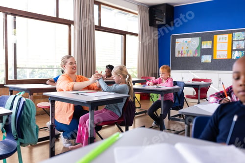 Preview: Female teacher assisting young girl with schoolwork in classroom, other diverse students studying