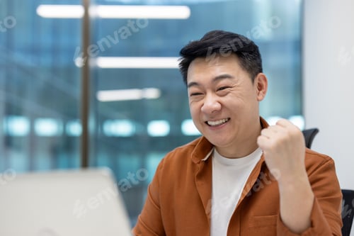 Preview: Cheerful Asian man in office celebrating success with fist pump while using laptop