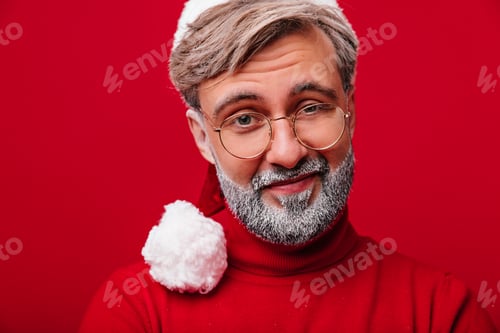 Preview: Curious grey-haired man in santa claus hat looking to camera. Indoor close-up shot of old male mode