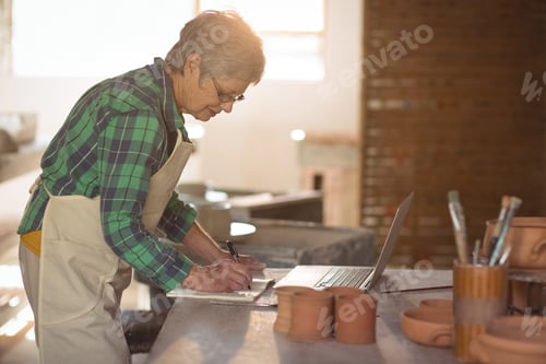 Preview: Female potter writing on a book while using laptop