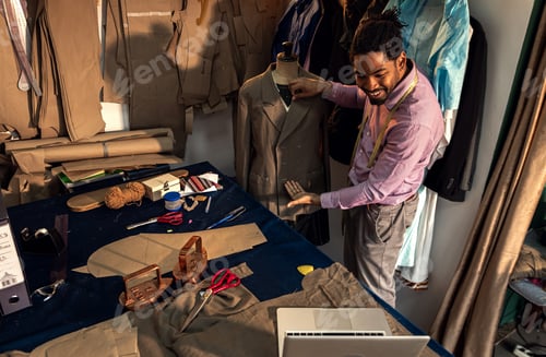 Preview: Male fashion tailor having video call with customer via laptop for the suit design in his workshop.