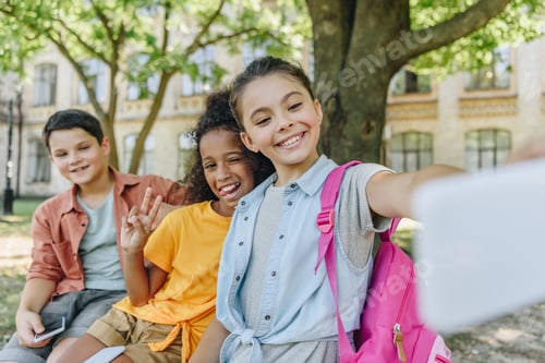 Preview: cheerful schoolgirl taking selfie with multicultural friends while sitting on bench in schoolyard