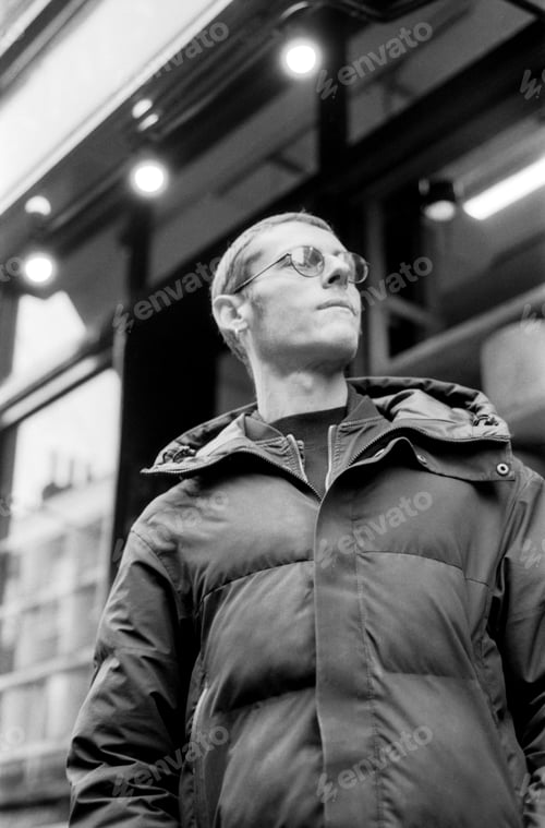 Preview: Young man wearing sunglasses and puffer jacket looking up in monochrome analog film style