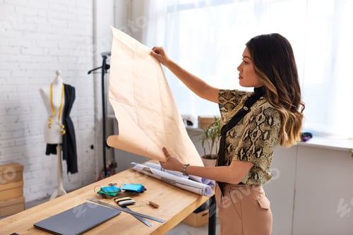 Preview: side view of young creative asian stylist looking at sewing patterns in personal fashion studio