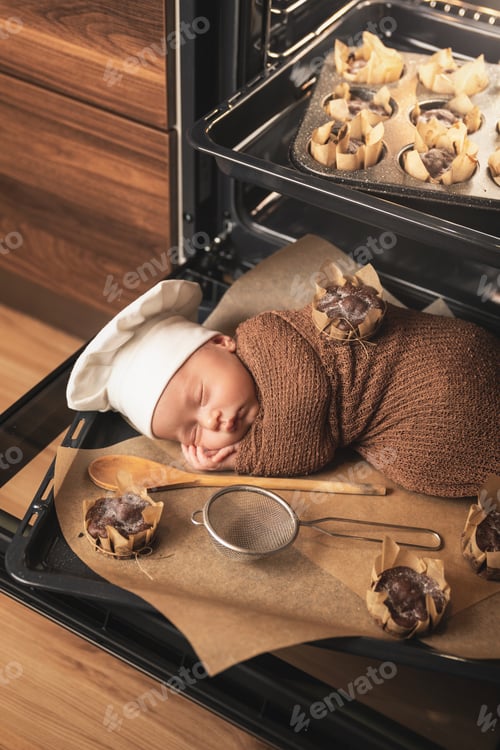 Preview: Newborn baby wearing chef's hat is lying on the oven tray with a muffins