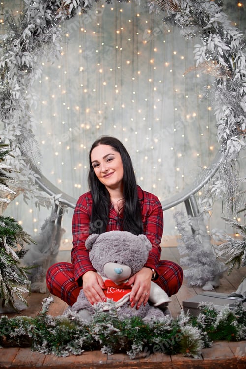 Preview: Young woman in the red pyjama hugs a teddy bear beside the Christmas decorated round swing